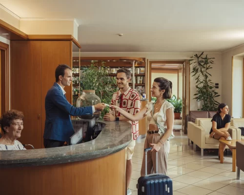 Ein Mann und eine Frau checken an der Hotelrezeption ein, w&auml;hrend eine Person im Hintergrund sitzt.