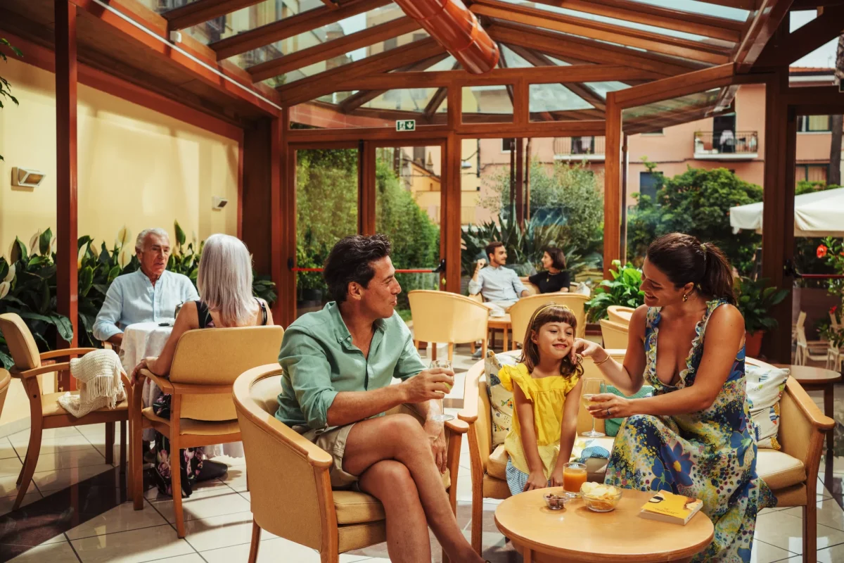Famiglia seduta in una luminosa sala interna con ampie finestre, mentre si godono bevande e conversazioni.